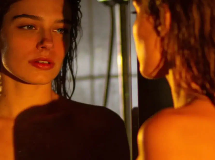 Primer plano cinematográfico de una mujer frente a un espejo con el cabello húmedo; su mirada es directa y reflexiva, iluminada por una luz lateral cálida que resalta la transparencia de sus ojos y la atmósfera íntima de la estancia.