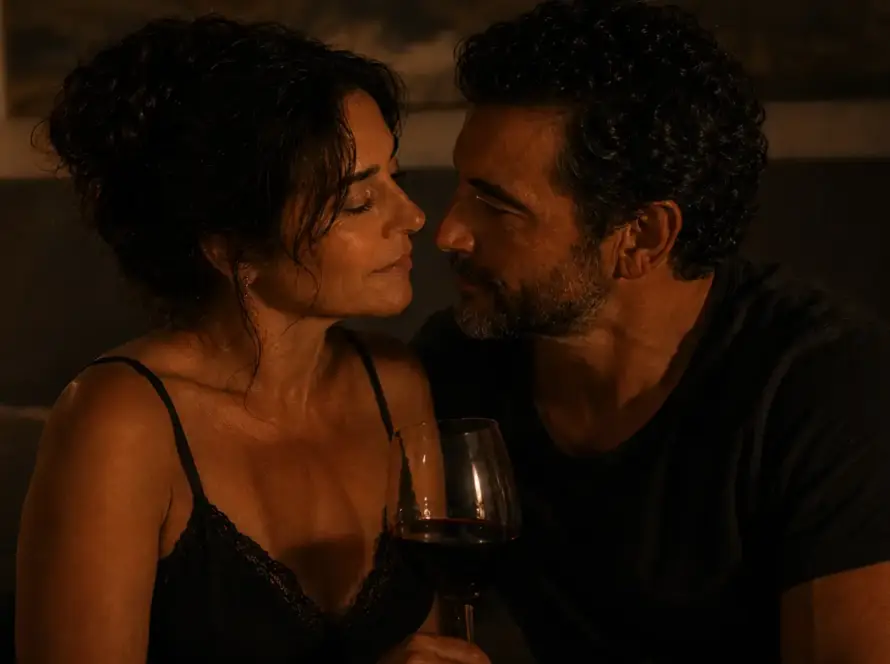 Isabel y Marcos sirviendo una copa de vino en la penumbra cálida del dormitorio, sin prisa, al comienzo de la noche — Serie Sexualidad Tántrica - Tántrika™